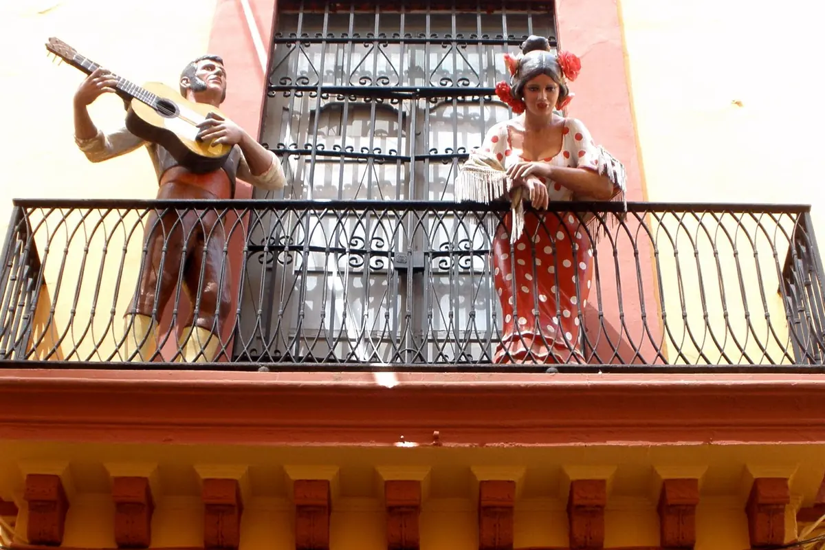 A painted guitarist and a flamenco dancer in a red polka-dot dress lean on a wrought-iron balcony on an ochre and red facade
