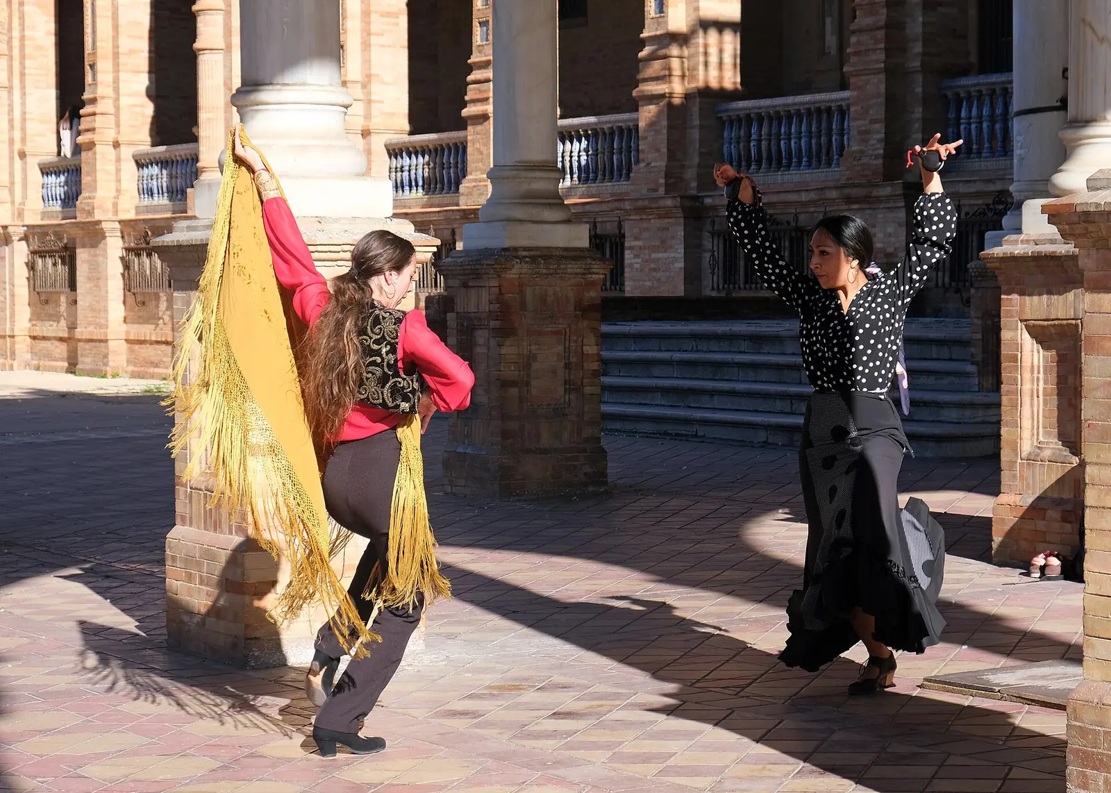 Two flamenco dancers, one with a golden shawl and one with castanets, dancing in a brick arcade at the Plaza de España in Seville